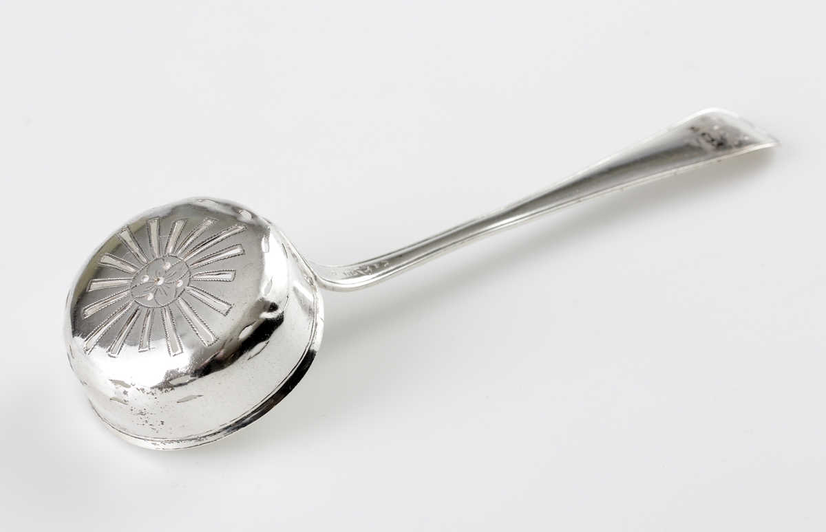 Strösked i silver.
Svensk trubbig modell. Perforerat runt skedblad med genombrutet strålformat mönster och fem runda hål. Längst ut två rader med avlånga genombrytningar. Ägarinitialer: "I.O.S." på baksidan av skaftet. Stämplar på baksidan av skaftet.