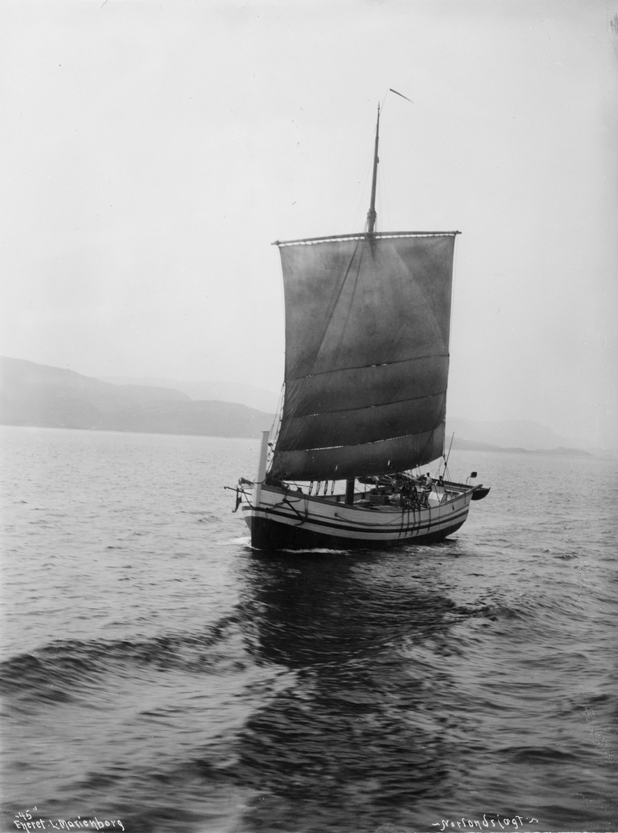 Motivet viser en nordlandsjekt for fulle seil i kystlandskap. To menn ombord, lettbåt henger i akterspeilet. Fotografiet er merket "45" Eneret L- Marienborg -Nordlandsjægt-.