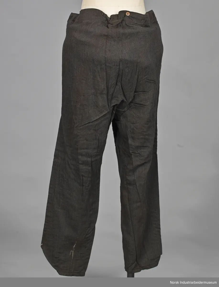 Bukser av vevet svart stoff. Lukkes med knapper foran. Den har metalknapper i linning og snor i livet. Buksen er foret med ternet stoff.