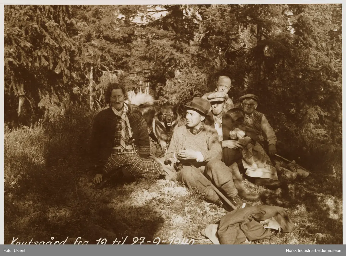 Fire menn, en dame og en hund sitter på mose i skogen
