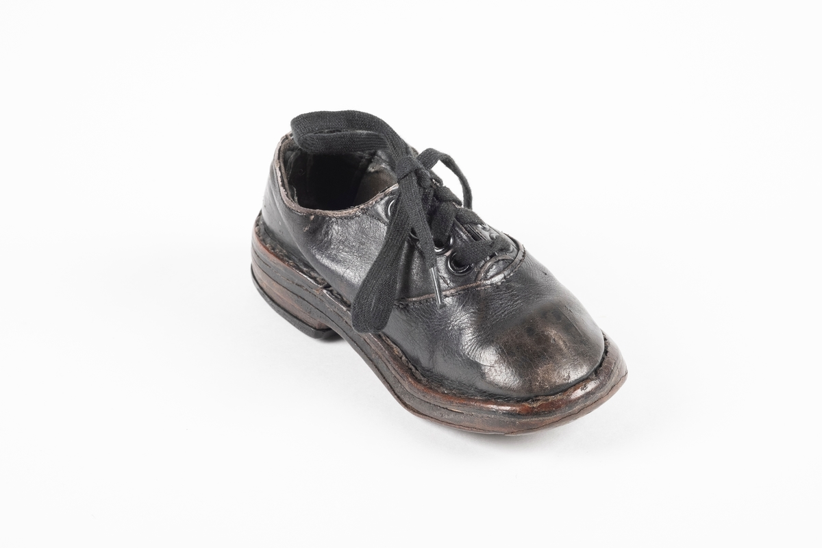 Et randsydd barnesko (venstre sko) av lær. Skoen har snøring med flate lisser. Maljene er av metall. Sålen er av lær og er forsterket med spiker på hælen og ved tuppen på undersiden.