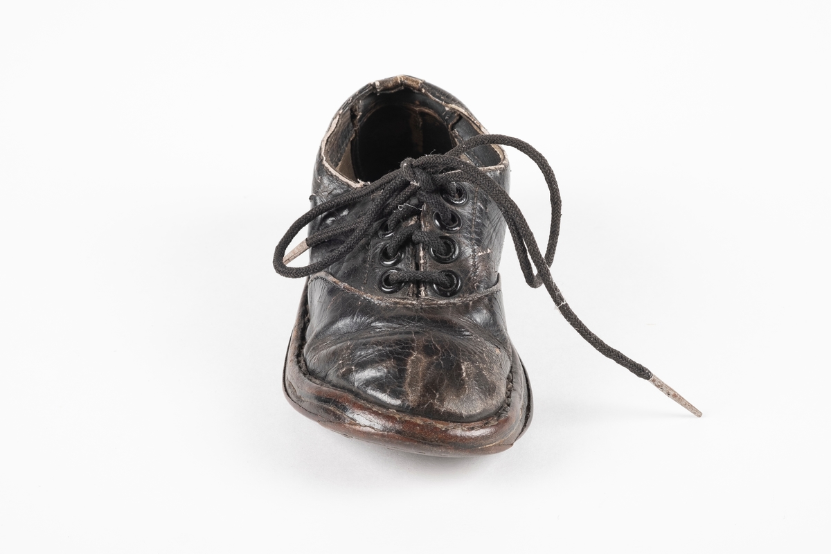 Et randsydd barnesko (høyre sko) av lær. Skoen har snøring med runde lisser. Maljene er av metall. Sålen er av lær og er forsterket med spiker på hælen og ved tuppen på undersiden.