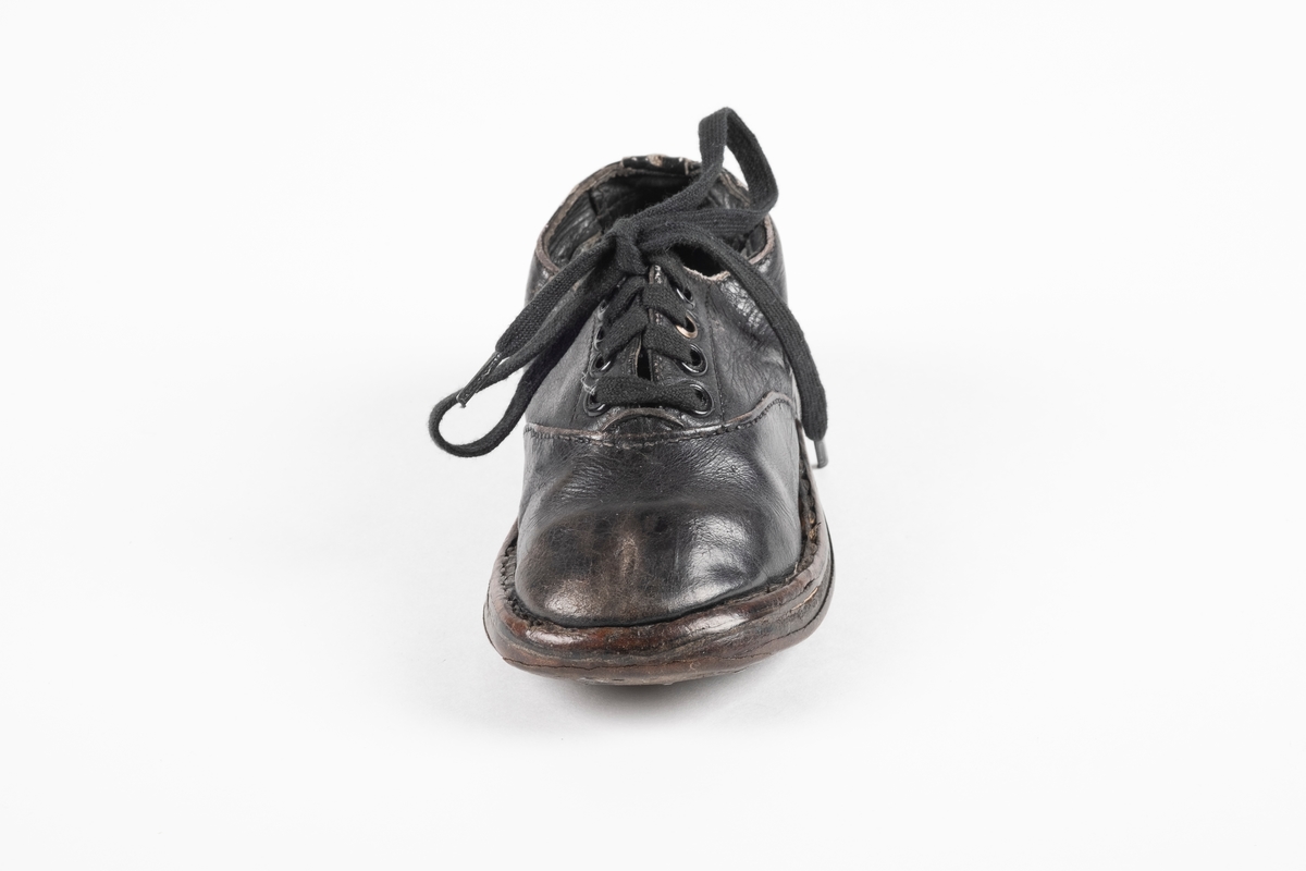 Et randsydd barnesko (venstre sko) av lær. Skoen har snøring med flate lisser. Maljene er av metall. Sålen er av lær og er forsterket med spiker på hælen og ved tuppen på undersiden.