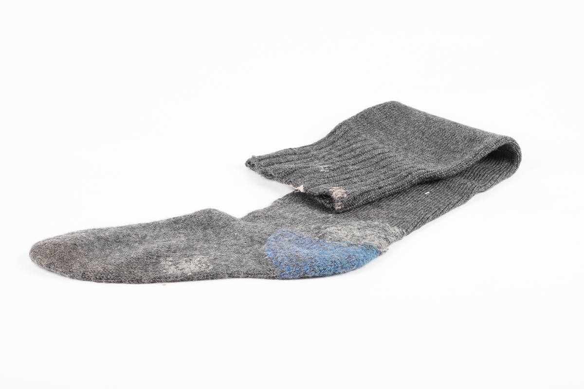 Ett par knestrømper strikket med grått ullgarn. Hælen er strikket med blått garn. På innsiden ved åpningen er det påsydd en lapp med fangenummer.
