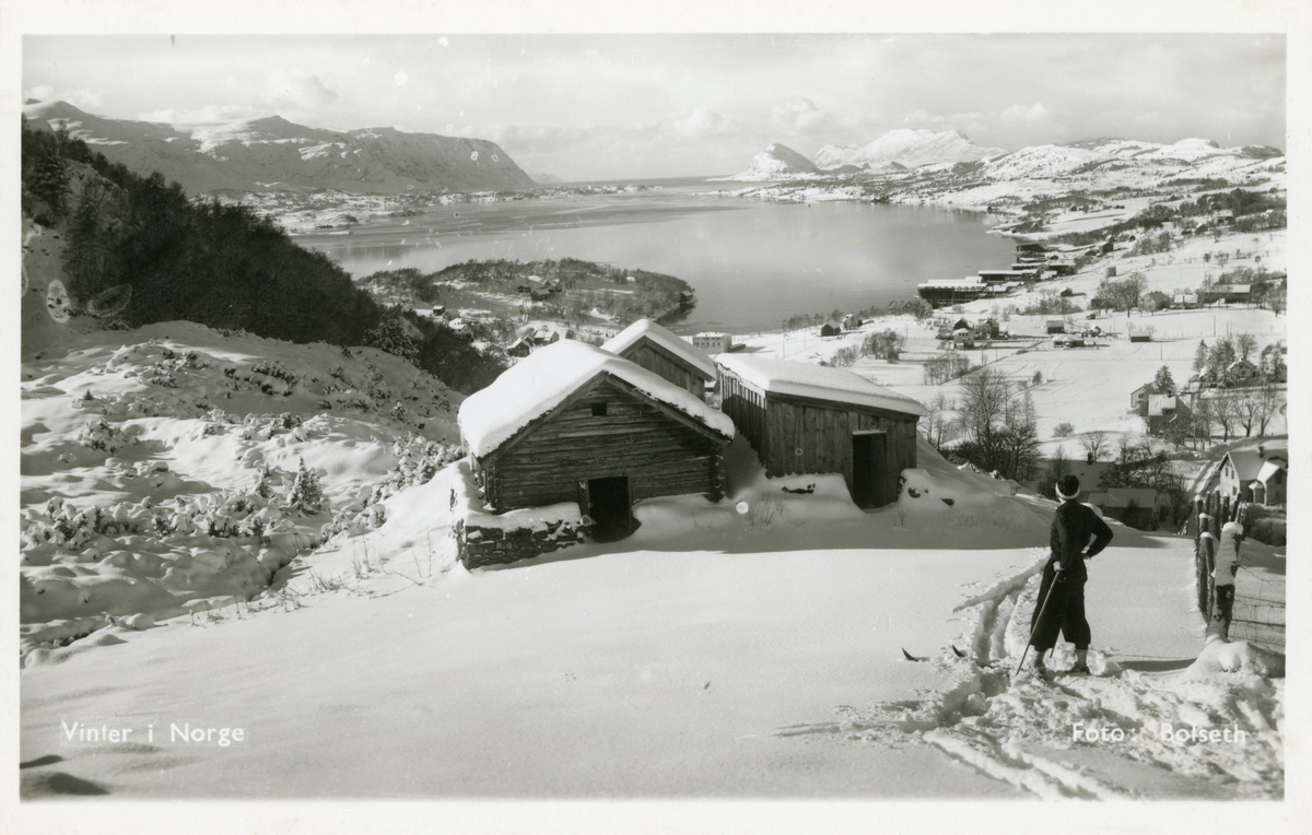 Motiv av noen gamle setershus og en skiløper i vinterdrakt. I bakgrunnen ses Spjelkavika og Åsestranda.
