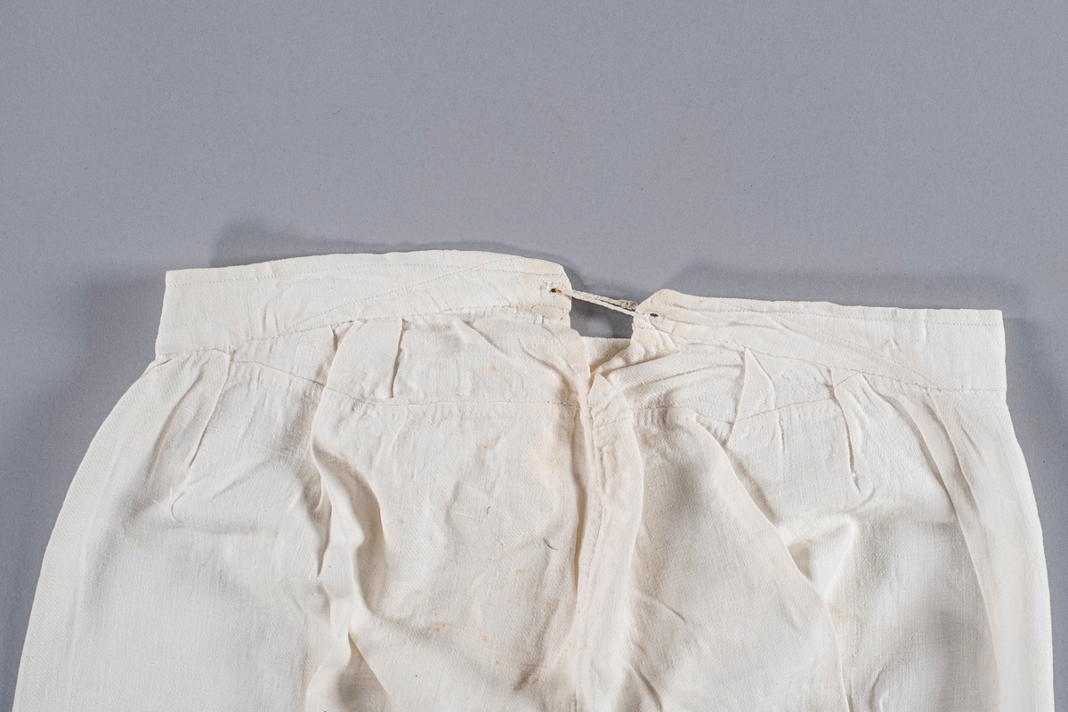 Underbukse i hvit bomull, åpning i front med to knapper, og åpning bak med snøring. Den har folder ved linningen. Nederst i beina er det splitt.