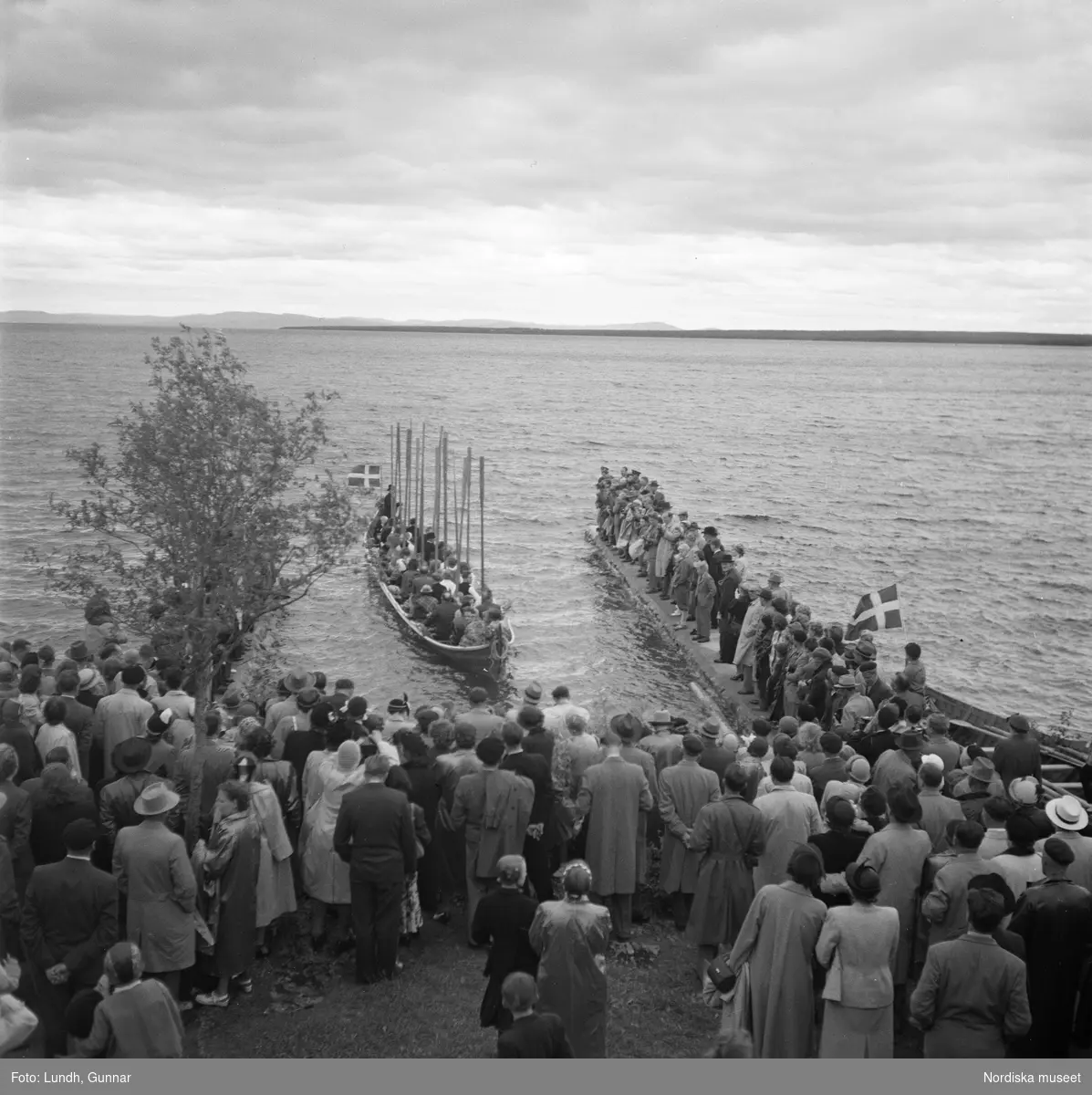 Motiv: (ingen anteckning) ;
En folksamling vid midsommarfirande, en folksamling står på stranden vid en sjö, kvinnor och män i folkdräkt ror en kyrkbåt.