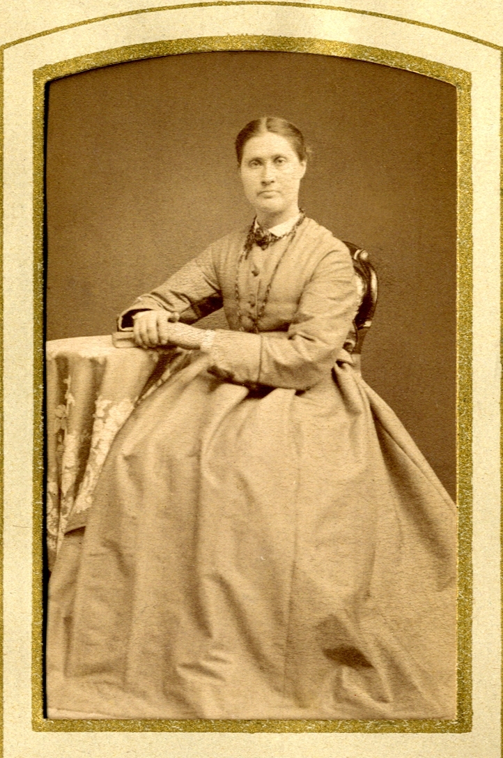 En kvinna i ljus klänning och liten vit krage, som sitter vid ett bord med lång duk. 
Helfigur, halvprofil. Ateljéfoto.