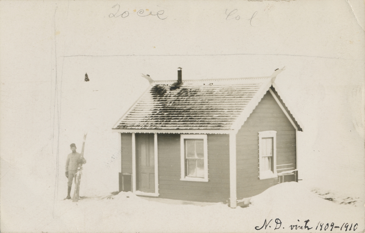 En mann med ski står ved et stovehus med dragehoder på endegavlene. Stova tilhørte Ingvald Skjeldrup og ble kallt "Haukelid"