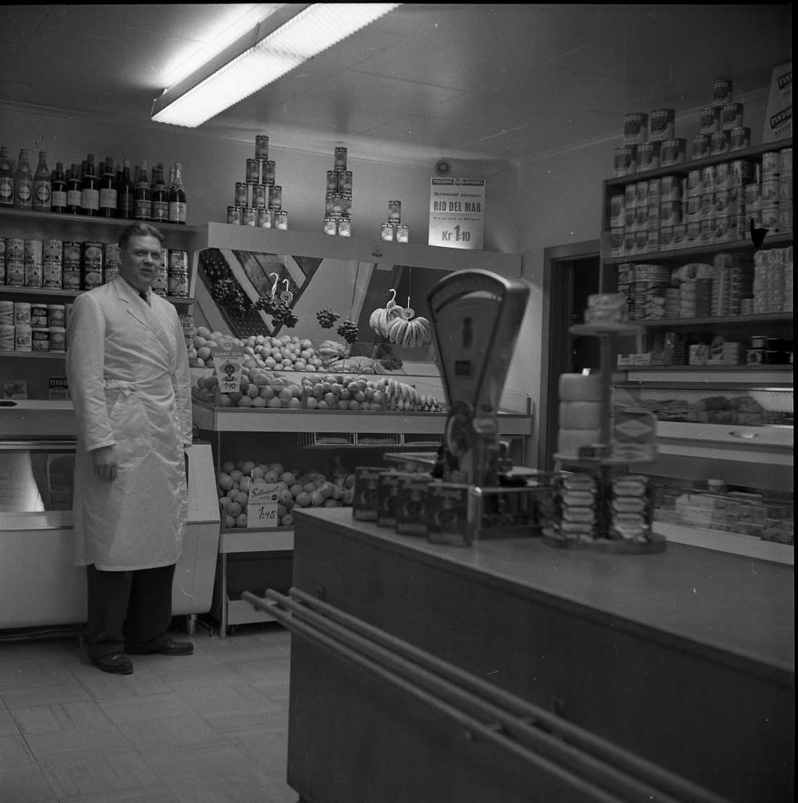 Köpman Åke Rahm i vit rock står i sin butik vid hyllor med konserver och disk med frukt.
