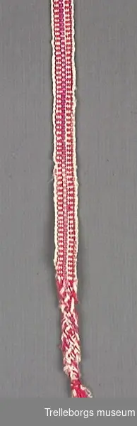 Vävt ullstrumpeband i vitt, rött, vinrött och lila. Ena änden är tvärt avklippt, den andra är flätad ca 8 cm och därefter ihopknuten.