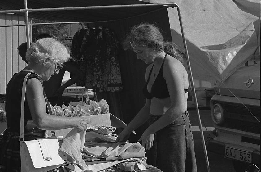 Marknad på okänd plats. Två kvinnor, sommarklädda, plockar med tyger på ett marknadsbord.