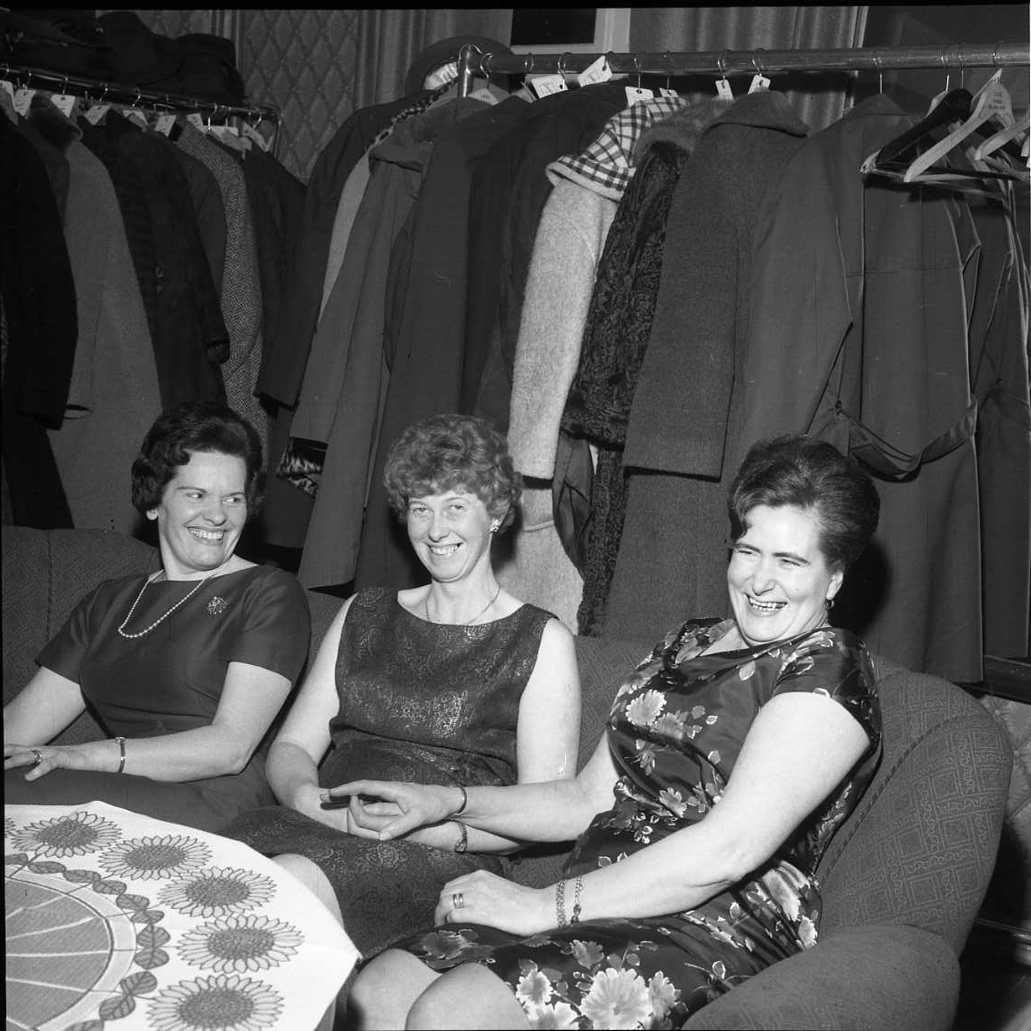 Tre festklädda kvinnor sitter i en soffa vid ett bord med duk och skrattar mot fotografen. Bakom dem står det klädställningar med ytterplagg. De befinner sig på Ribbagården i samband med årsmöte för Gränna AIS.