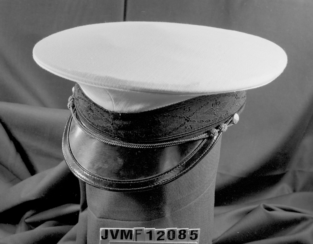 Skärmmössa med vitt sommarkapell. Svart mössband med invävt mönster av krönt bevingat hjul och SJ. Gyllene stormträns, och gyllene mösstränsknappar (1953)
Broderat mössmärke.