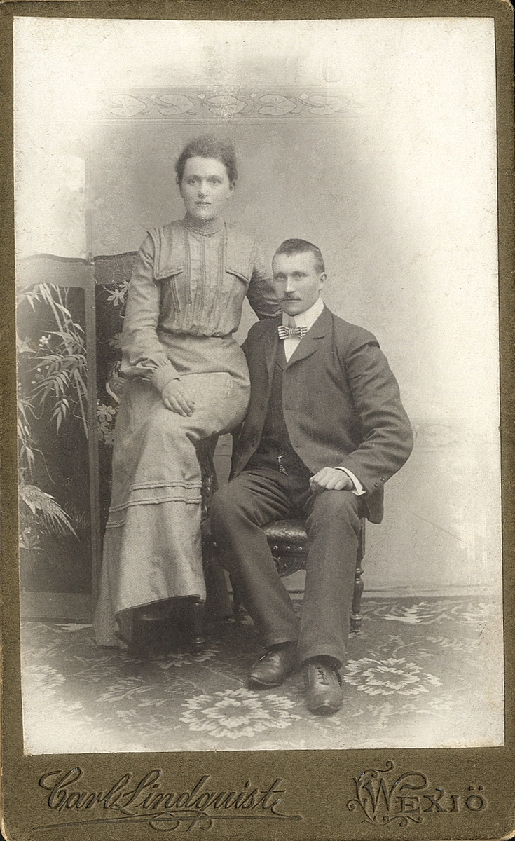 Foto av ett okänt par. Hon är klädd i klänning med hög krage. Han är klädd i kavajkostym med väst, klockkedja och stärkkrage med randig fluga.
Helfigur. Ateljéfoto.
