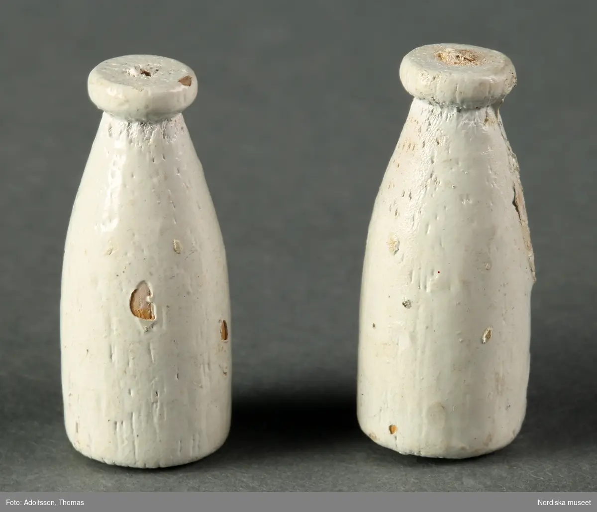 Två vitmålade mjölkflaskor av trä. Hör till dockskåpsinredningen i matkällaren till dockskåp NM.0331721+.
