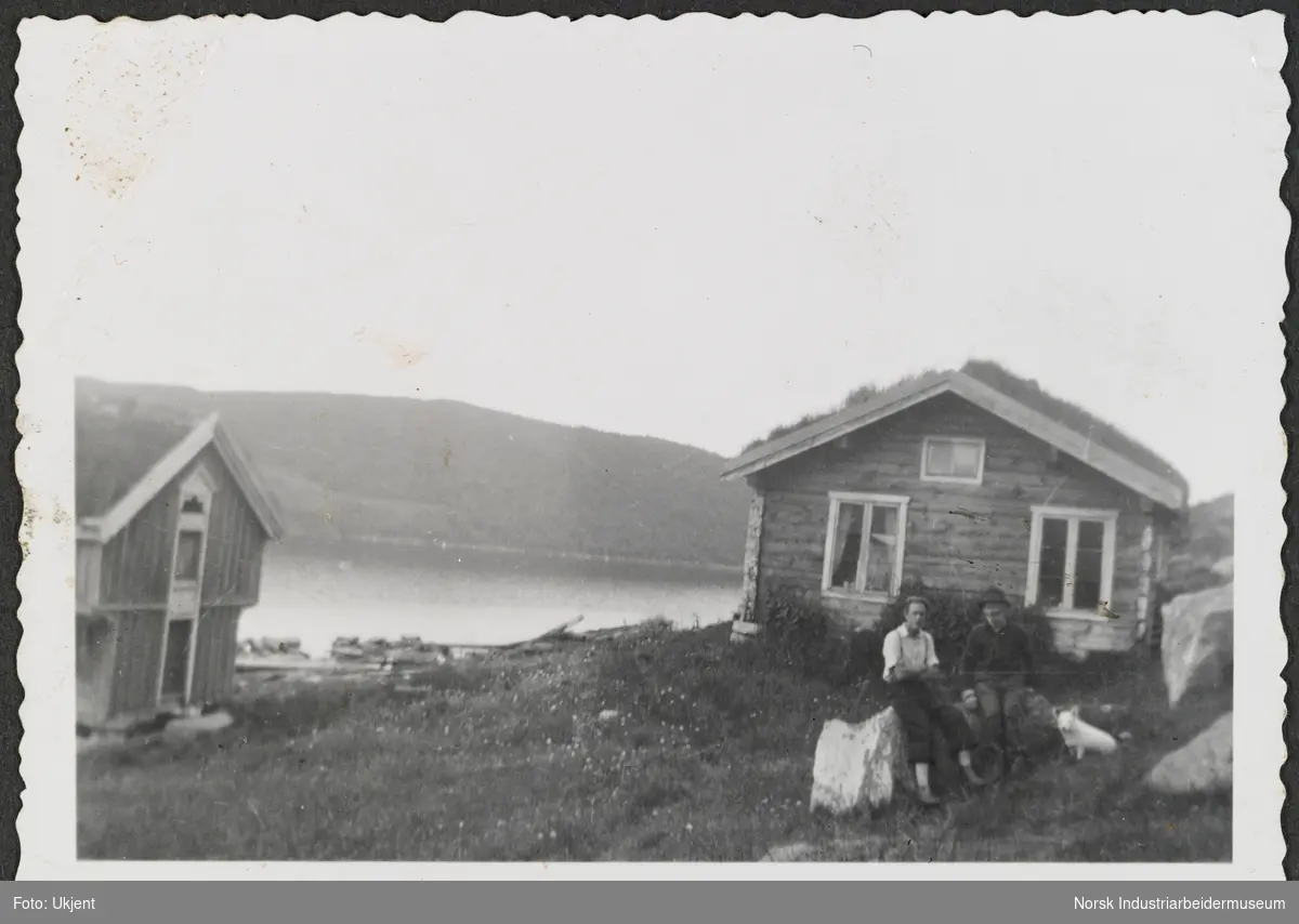 To menn og hund sitter på stein foran bolig og gård på Møsstrond. Møsvatn i bakgrunn