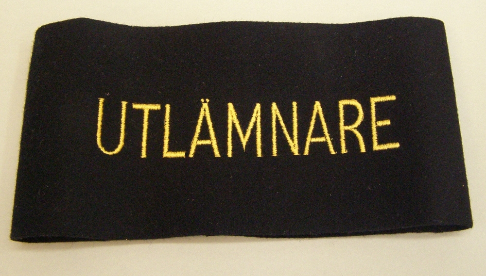 Armbindel av svart kläde med texten UTLÄMNARE broderat i gult. Bokstäverna är 27 mm höga.
