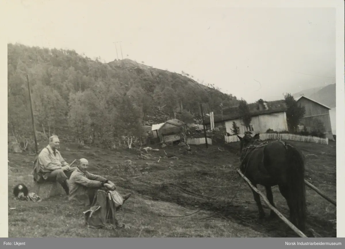 Vetle Skinnarland og en mann sitter i lia på gården Skinnarland. Foran står hest iført seletøy og kjerre. I bakgrunn sees våningshus, låve og uthus på gården