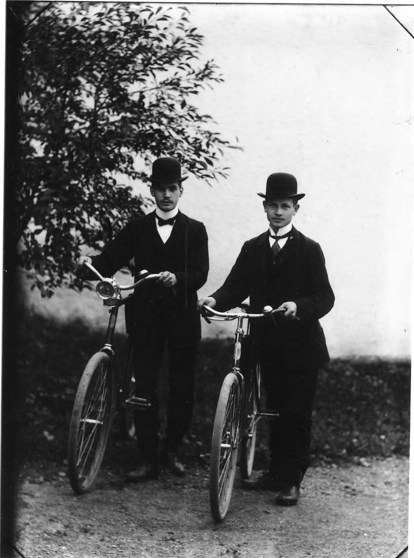 Gruppbild med två unga män med cyklar. De är finklädda med fluga och hatt. Mannen till vänster är Ludvig Roman.