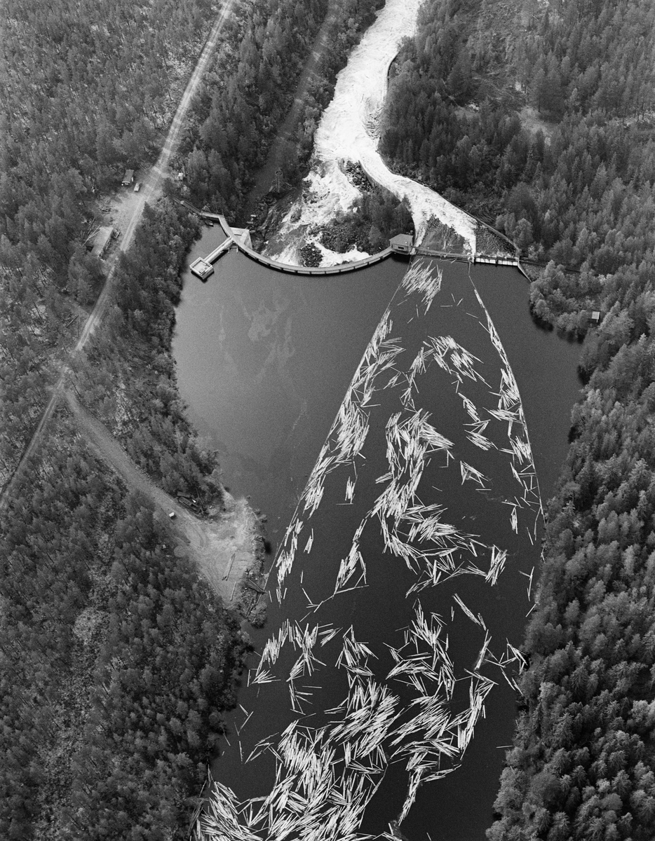 Flyfotografi tatt over kraftverksdammen i Osfallet i elva Søndre Osa i Åmot i Hedmark i mai 1984, den siste sesongen det foregikk tømmerfløting i denne delen av Glomma-vassdraget.  Fotografiet viser hvordan tømmerstokkene kom flytende med strømretningen over kraftverksdammen, der det var lagt ut lenser for å lede virket mot tømmerløpet som var plassert mot det nordre landet, hvor fallhøyden var mindre enn nedenfor den buete hoveddammen mot søndre landside.  Damanlegget på fotografiet skal ha vært ferdigstilt i 1947.  Den ble noe ombygd i 1981.  Ettersom vassdragslovgivinga påla dem som drev tømmerfløting å fjerne installasjoner i vann ved opphør av virksomheten, ble tømmerløpet ved Osfallsdammen revet umiddelbart etter at fløtinga på dette fotografiet var avviklet.  Energiproduksjonen ved denne lokaliteten foregår i en kraftstasjonsbygning fra 1914, cirka en kilometer nedenfor den avbildete dammen, hvor det er installert to francisturbiner som kan yte 5.5 megawatt.  Den opprinnelige dammen ble ødelagt ved et dambrudd i 1916.  Det nåværende kraftverket utnytter et fall på 41 meter i elva Søndre Osa.  Vegen på sørsida av vassdraget fører ned til kraftstasjonen og derfra mot Fylkesveg 215 ved Oset.