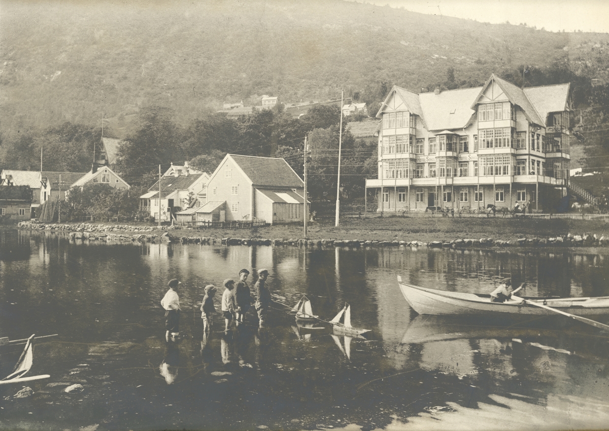 Motiv fra Sjøholt med naustrekke og hotellet til høyre. I forgrunnen står noen gutter og leker med båt i vannet.