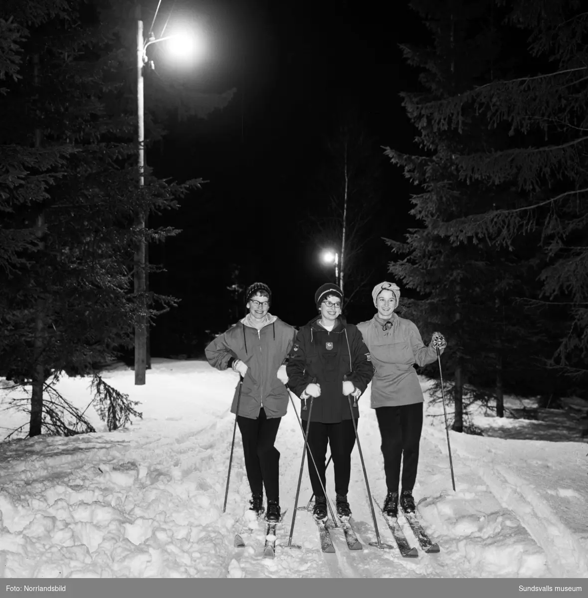 Tre kvinnor åker skidor i elljusspår. Möjligen på Södra berget.
