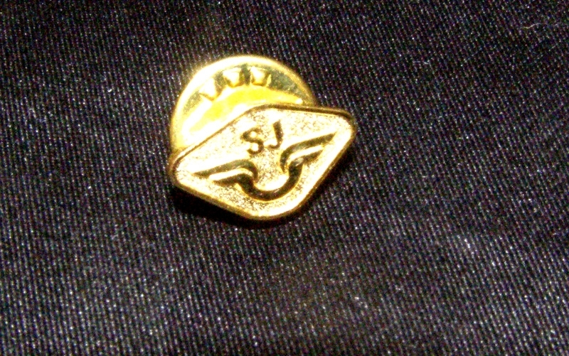 Rombformad pin av metall med SJ:s logotyp. Fjärilsklämma på baksidan för fastsättning i kläderna.