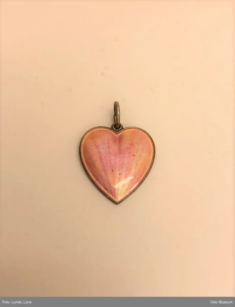 Anheng i form av et hjerte, forsiden er belagt med rosa emalje. "Fadervår" er inngravert på smykkets bakside.