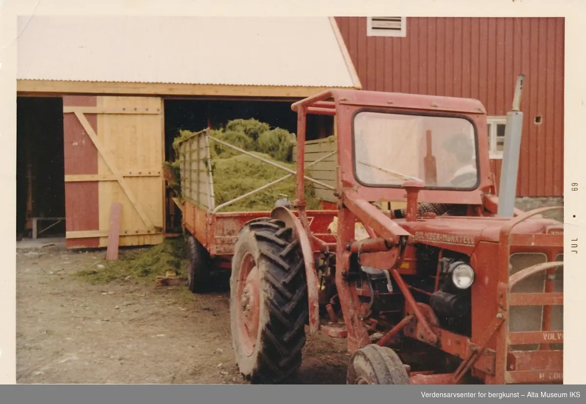 Rød traktor med tilhenger og høy foran låve