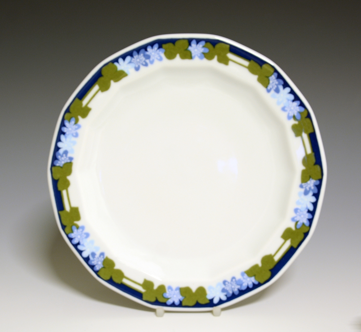 Mangekantet tallerken av porselen med hvit glasur. Dekorert med blåveisdekor. 
Modell: Octavia, tegnet av Grete Rønning i 1977.
Dekor: Blå Anemone