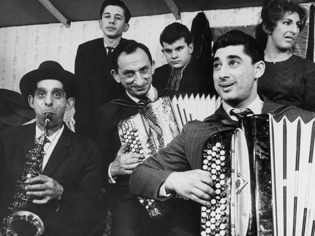 En grupp romer spelar musicerar i en bostadsbarack. En man spelar saxofon och två andra spelar dragspel.