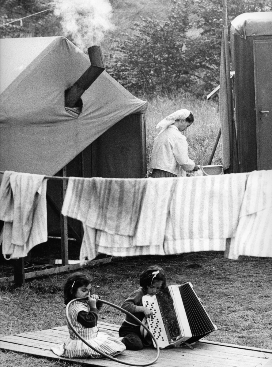 Två barn sitter på ett brädgolv på marken. Bakom dem hänger tvätt och en kvinna står vid ett tält och ordnar med något.