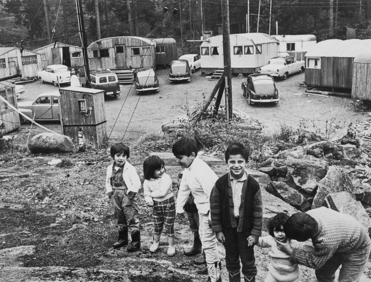 En samling barn står uppställda för fotografering i ett av de läger som Stockholm stad upprättade för romer i slutet av 1950-talet. I bakgrunden syns husvagnar och vagnar samt tält.