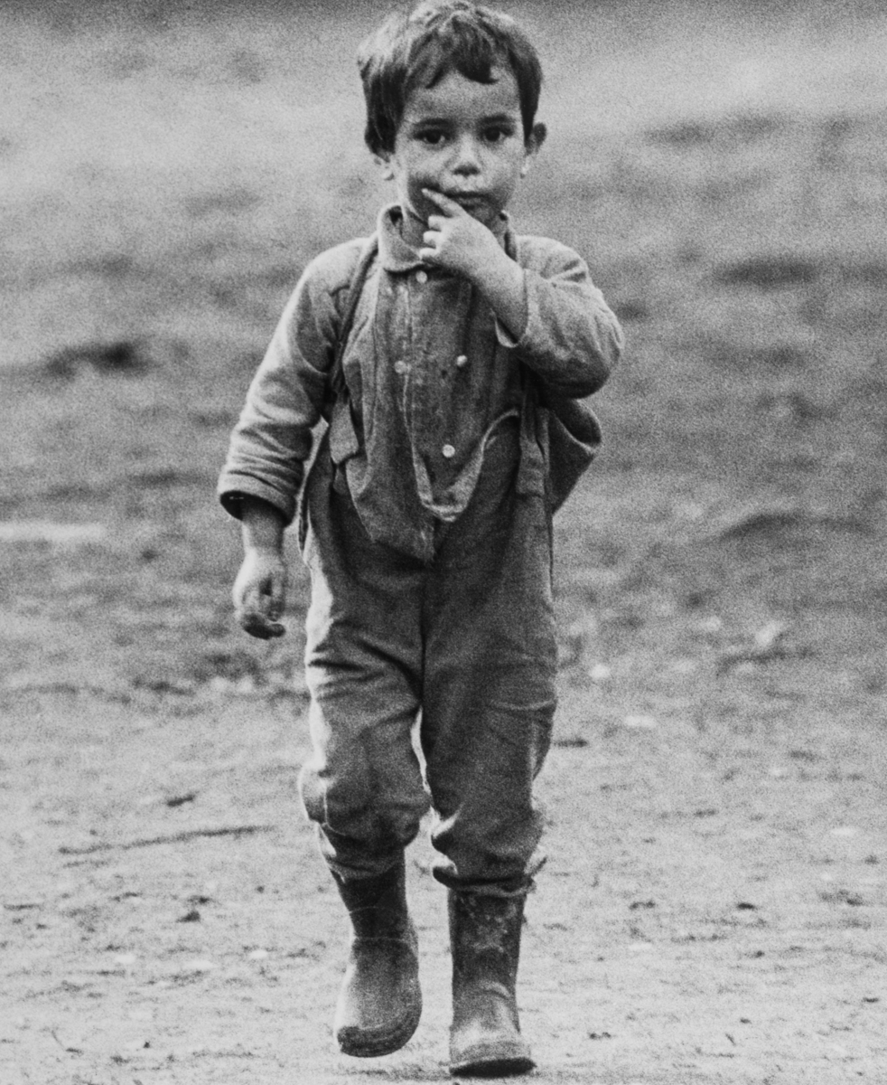 En pojke går på en plan yta. Han bär hängselbyxor och gummistövlar.