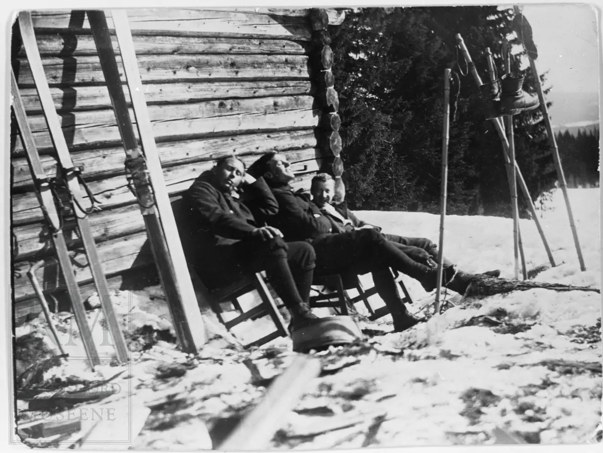 En gruppe menn står og sitter utenfor en tømmerbygning. Ski oppetter veggen og staver i snøen.