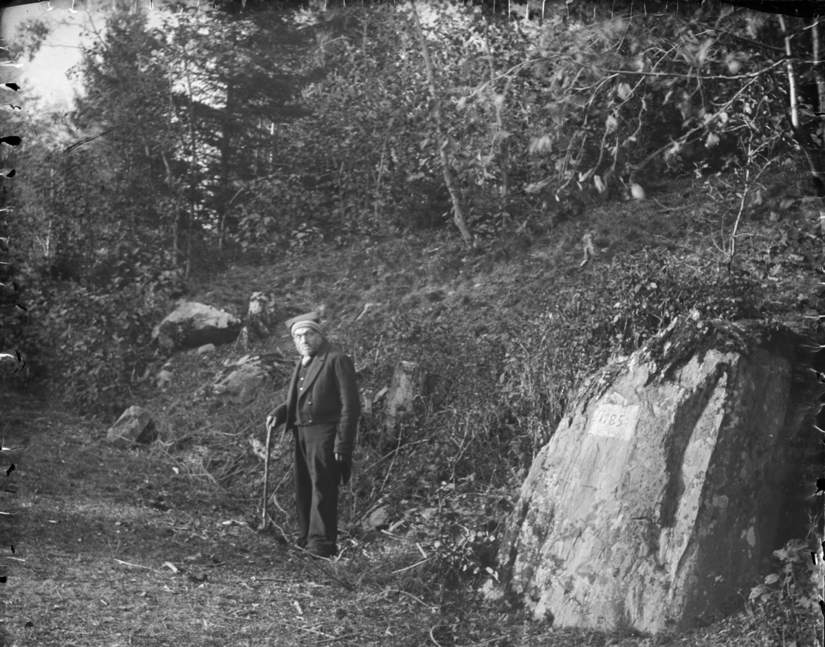 En stor stein ved et skogholt, nylig gravd fram, med påskriften 1785. I bakgrunnen en mann i vadmelsdress, raudlue og stokk.