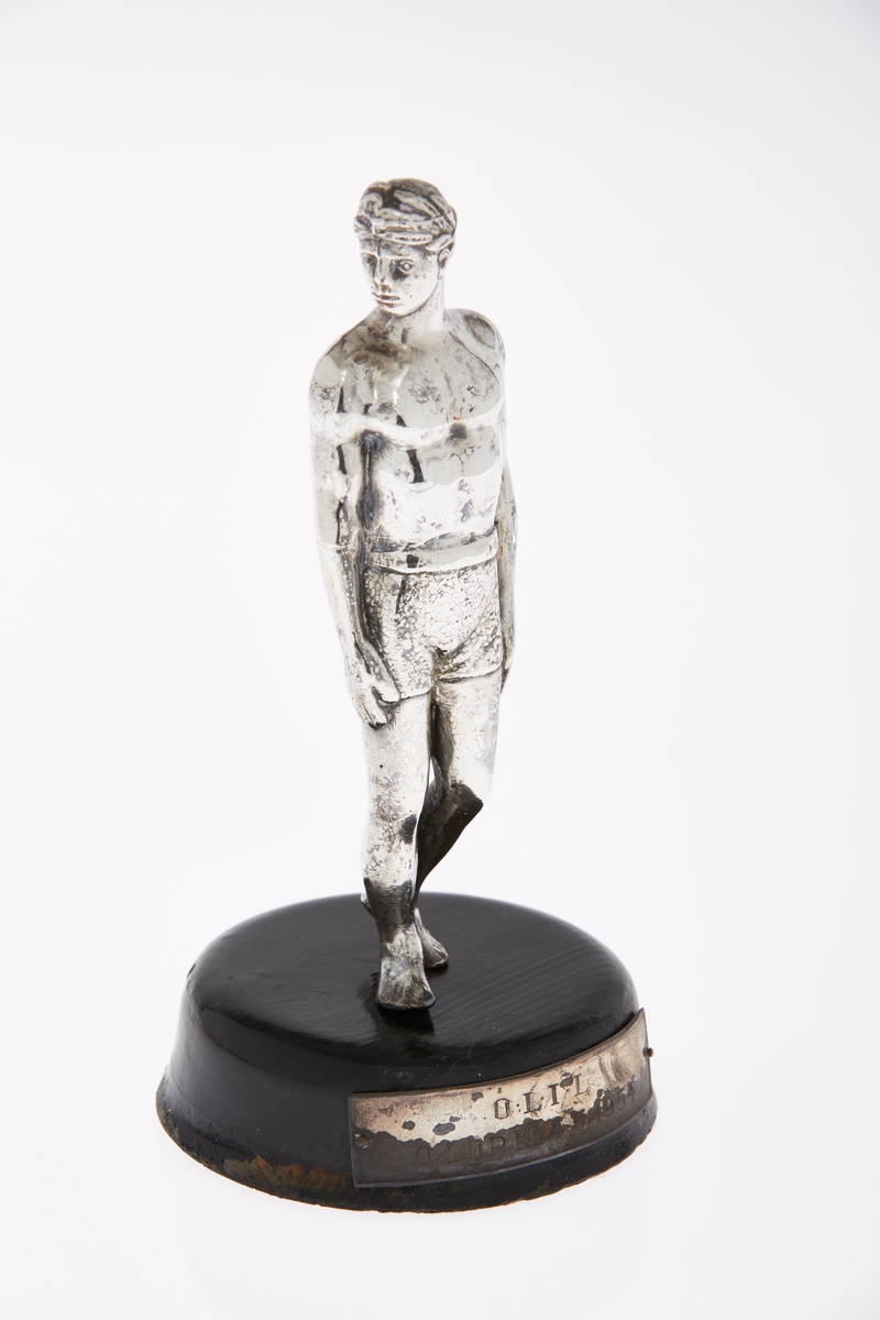 Statuett i sølvfarget metall, en atlet, med støtte i tre. Plakett på trestøtten med inskripsjon.