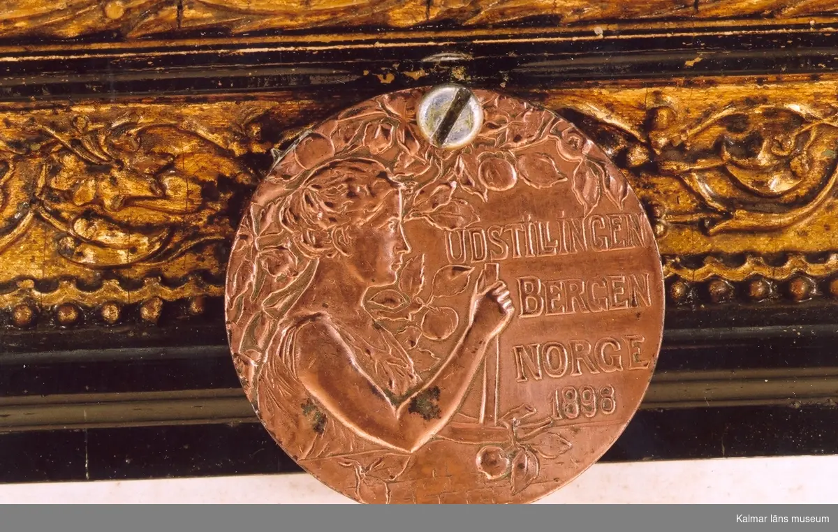 KLM 39586:54 Medalj av metall och tillhörande ask av trä, papp och textil. Medaljen är monterad på diplomets ram (KLM 39586:60). Bronsmedaljen tilldelades Gustaf Axteliu, vid Udstillingen Bergen, Norge 1898.