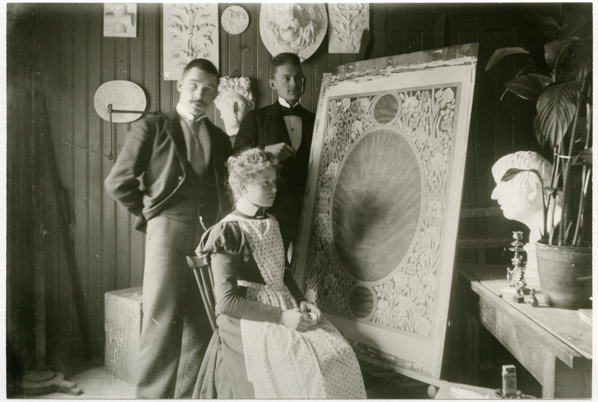 Stockholm. 
Kvinna sittande framför målning på staffli. Två män står bakom. 1901.
