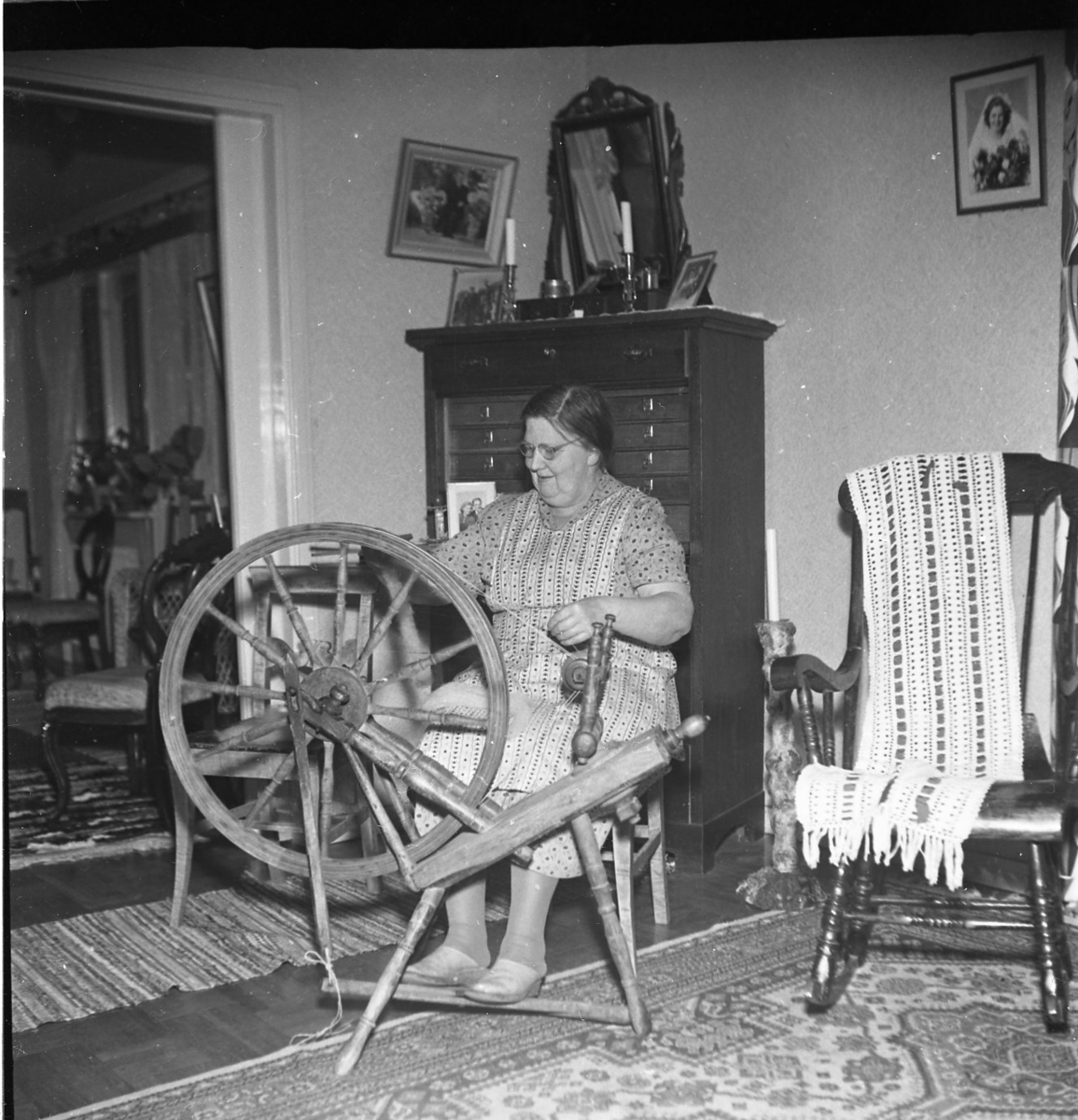 Fru Rut Sääv arbetar vid sin spinnrock i sitt hem. Bakom henne står en hög sekretär med en pigtittare ovanpå. Till höger en gungstol med troligen en virkad genombruten sjal