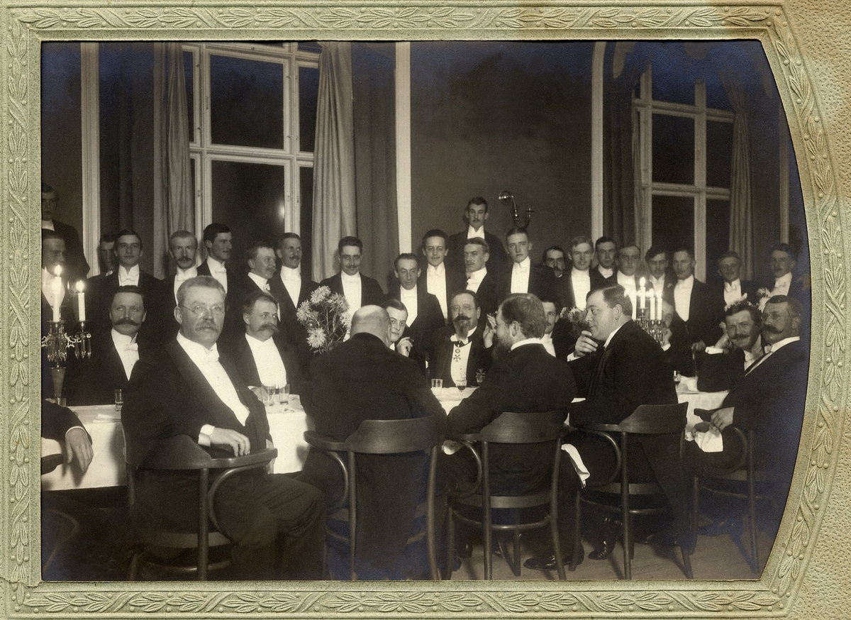 En sammankomst med frackklädda anonyma herrar (enbart), samlade vid några middagsbord med kandelabrar 
m.m.
Trol. någon akademisk sammankomst i Uppsala.