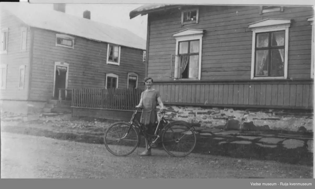 Dagmar Sundelin med sykkel foran Hallonengården med badstubygning til høyre. Vadsø ca 1930- 1940