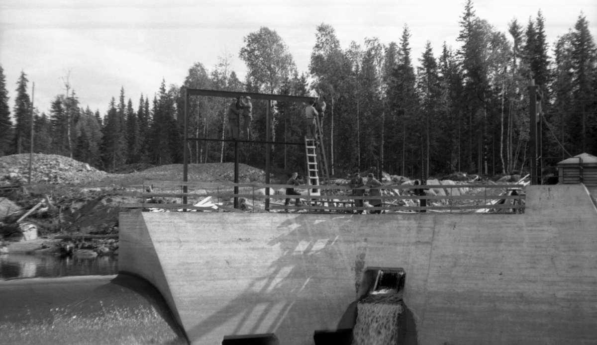 Dammen ved Kvernfallet i elva Søndre Osa i Åmot i Hedmark, fotografert i motstrøms retning i 1941, På dette tidspunktet var dette anlegget, som ble satt i drift i 1936, ennå forholdsvis nytt, og i bakgrunnen på bildet ser vi fortsatt jord- og grushauger fra anleggsperioden. Hovedmotivet her er imidlertid damkonstruksjonen, som var utført i armert betong. Til venstre i bildet ser vi hvordan vannet rant over overløpsdelen, som hadde avrundet krone. I midten og til høyre på bildet ser vi en mer kantete mur som omsluttet det bassenget der turbinrøret hadde sitt innløp. Det skulle føre driftsvann ned til kraftstasjonen ved Brufallet, noen hundrede meter lengre nede ved elveløpet. Turbinrøret vises ikke på dette fotografiet, men vi ser tre noenlunde kvadratiske åpninger i dammuren, to helt nederst og en litt høyere oppe. Gjennom den sistnevnte strømmet det vann da dette bildet ble tatt. Disse åpningene skulle antakelig brukes som avløpsventiler når det var vel mye vann i dammen. De kunne åpnes og lukkes ved hjelp av hev- og senkbare luker på et stålstativ som var montert på motstrøms side av dammuren. Da dette fotografiet ble tatt arbeidet to menn med dette stativet fra stiger som sto på damkrona, mens to andre sto i stativet, uten stigeforbindelse nedover. Tre andre sto ved rekkverket på damkrona.