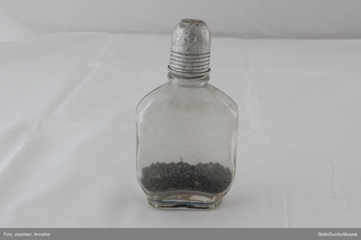 Glassflaske (lommelerke) som inneholder hagl, ammunisjon, for å lade patroner selv. Glasset i flasken er ujevn (bølget). Lokket av metall er formet som et lite beger med synlige gjenger utenpå.