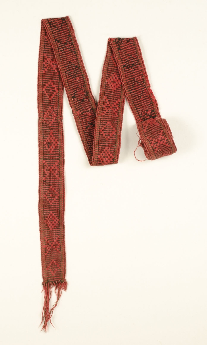 Pälsband, mönstervävt av rött och svart ullgarn. Frans av varpen, knuten. Skarvat på två ställen. Invävt årtal: 1848