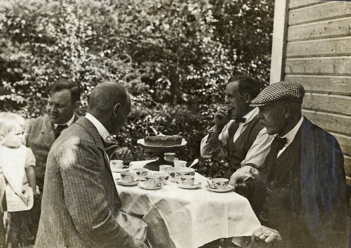 Fyra herrar och en liten flicka sitter runt ett kaffebord utomhus.
