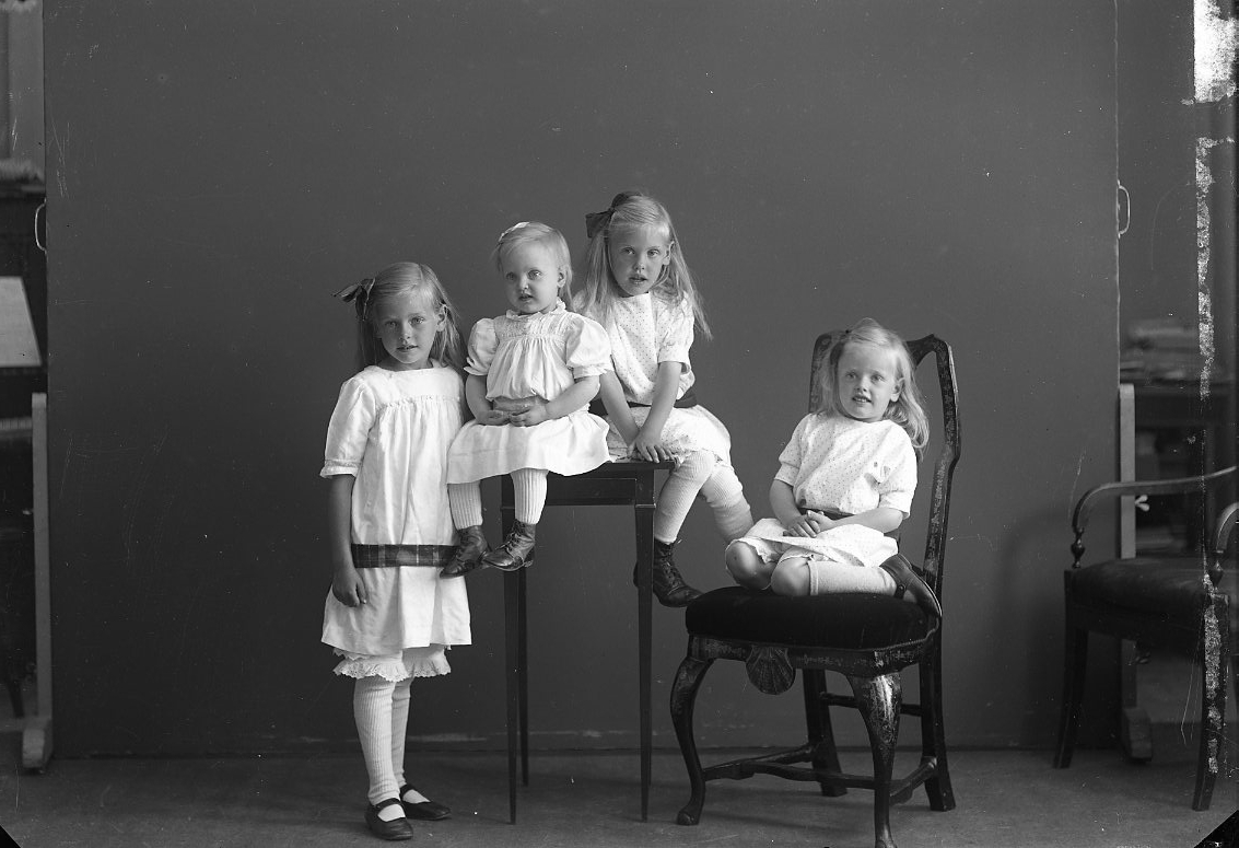 Troligen en syskongrupp. Fyra flickor som står, sitter på ett litet bord och på en stol. Gissningsvis mellan 1 och 6 år. De äldre flickorna har rosett i håret.