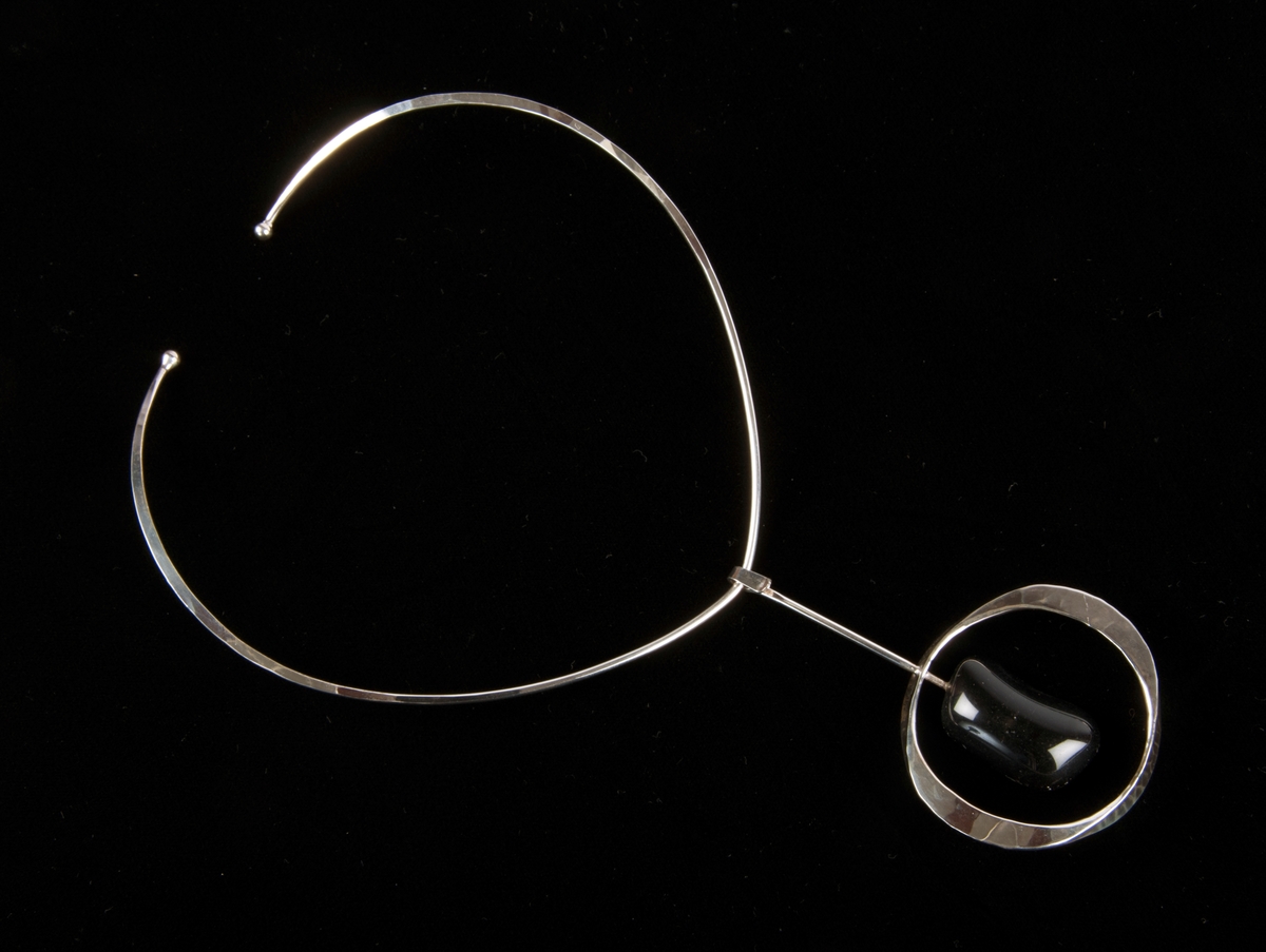 Anheng: Åpen, flathamret ring. I ringen er det tredd en sølvstang med bærekrok til kjede i den ene enden. I den andre enden er festet en stein. Sotgrå trapesformet naturstein. Ringen er hamret i to plan, 2+2 sider.