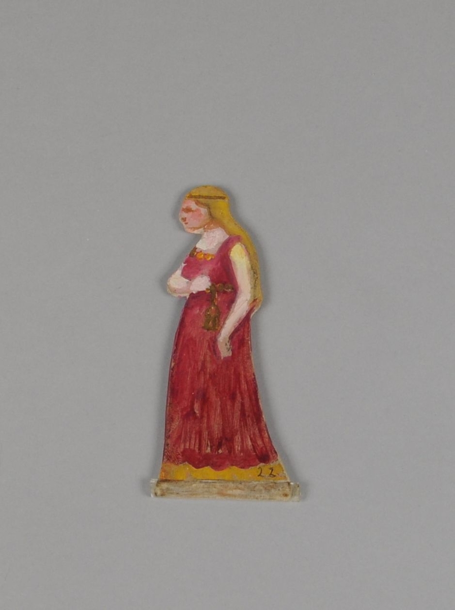 Figur av papp. Forestiller dame i sideprofil, med rød kjole og gullfarget belte. Figuren er malt på begge sider.